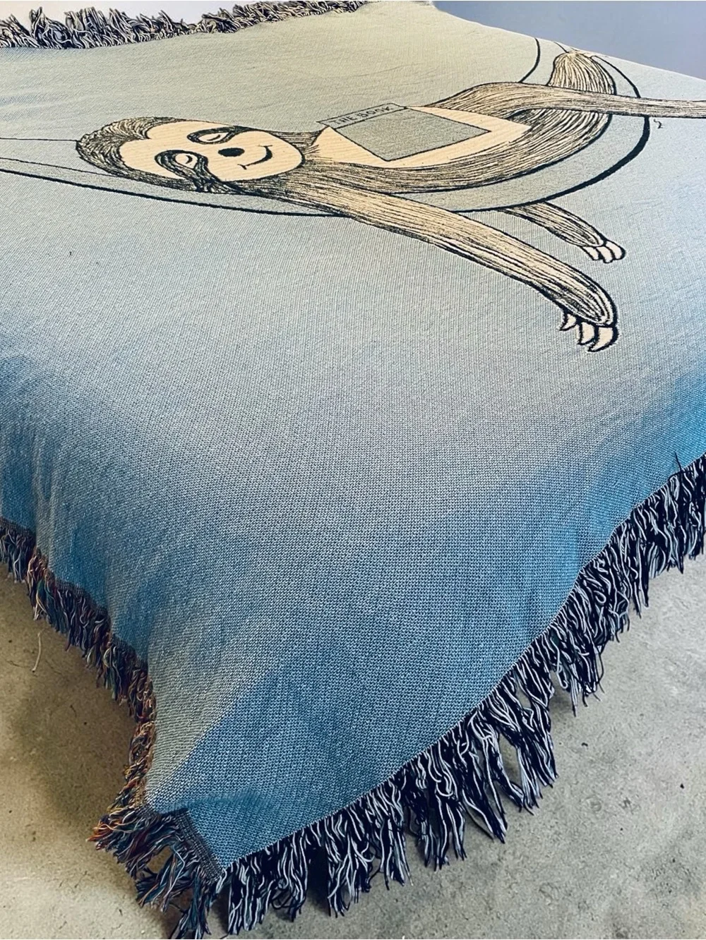 Outcasty cotton throw, relaxing sloth with book, shown on a Queen size bed